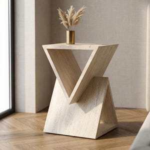 May include: A light-coloured, geometric side table with a triangular design. The table has a natural stone appearance and features an open, inverted V-shape. A small gold vase with dried plants sits on top.