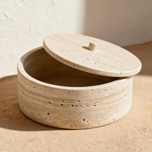 Pode incluir: Um recipiente redondo de pedra travertino bege com uma tampa circular a condizer. A superfície natural porosa apresenta faixas horizontais e um pequeno puxador cilíndrico centrado na tampa. O recipiente tem uma parede espessa.
