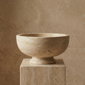 Pode incluir: Uma tigela de pedra bege com formato arredondado, colocada sobre um pedestal quadrado correspondente. A tigela tem uma abertura larga e uma superfície texturizada. O pedestal também é de pedra.
