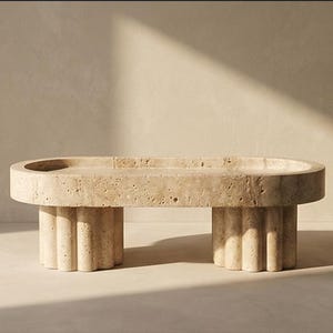 May include: A beige, oval coffee table with a textured surface. It features two fluted column-style legs. The design is minimalist and modern, suitable for living room decor.