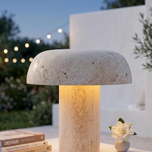 May include: A mushroom-shaped table lamp crafted from beige travertine stone with a porous, textured surface. The lamp features a wide, rounded shade resting on a cylindrical base, emitting a warm, soft glow from the center of the structure.