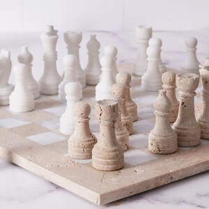 May include: A chess set featuring white and beige chess pieces on a beige and white chequered board. The pieces include pawns, rooks, bishops, knights, a queen, and a king. The set is made of stone.