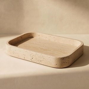 May include: A rectangular tray crafted from beige travertine stone, featuring a porous, textured surface with natural pits and variations. The tray has rounded corners and raised edges, showcasing the organic, earthy tones of the natural stone.