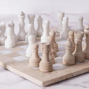 May include: A complete chess set with white and tan pieces on a beige and white checkered board. The chess pieces are carved with detailed designs. The board is made of stone.