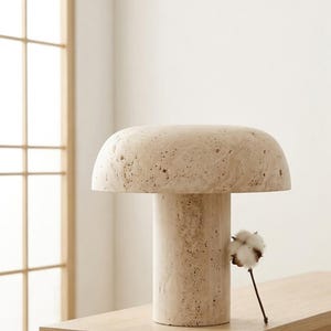 May include: A mushroom-shaped table lamp crafted from beige, porous travertine stone. The lamp features a wide, rounded dome top resting on a cylindrical base, showcasing the natural, textured, and pitted surface characteristic of the stone material.