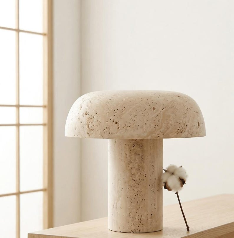 May include: A mushroom-shaped table lamp crafted from beige, porous travertine stone. The lamp features a wide, rounded dome top resting on a cylindrical base, showcasing the natural, textured, and pitted surface characteristic of the stone material.