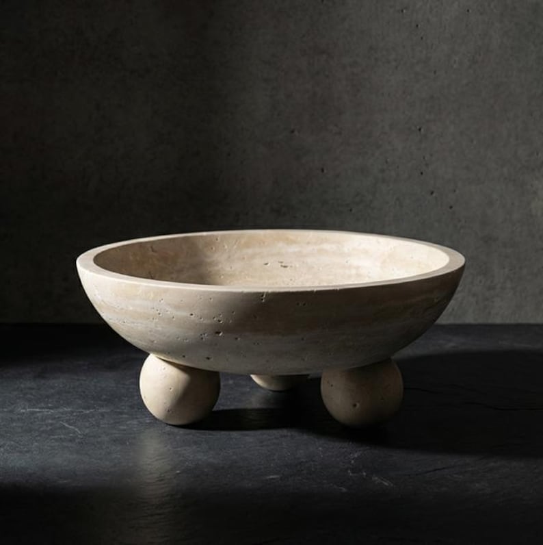 May include: A beige, round bowl with a textured surface, supported by three spherical feet. The bowl has a wide, shallow design and is made of a stone-like material. The neutral color and simple design make it a versatile decorative piece.