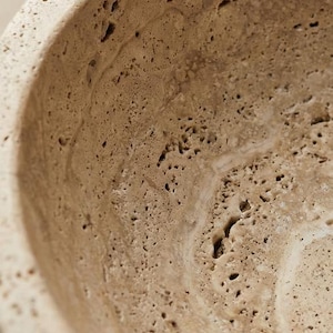 May include: Close-up of a beige, textured bowl with a porous surface. The bowl's interior has a smooth, curved design, and the material appears to be stone or a similar natural substance. The bowl is likely for decorative or functional use.