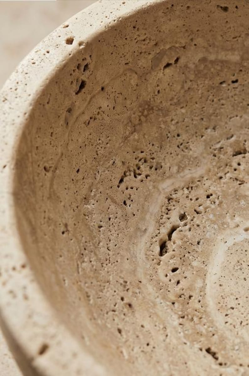 May include: Close-up of a beige, textured bowl with a porous surface. The bowl's interior has a smooth, curved design, and the material appears to be stone or a similar natural substance. The bowl is likely for decorative or functional use.