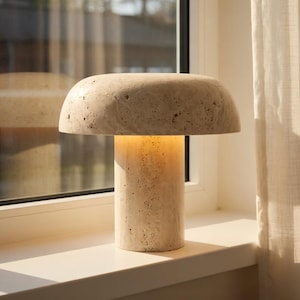 May include: A mushroom-shaped table lamp crafted from beige travertine stone with a porous, textured surface. The lamp features a wide, rounded dome shade resting on a cylindrical base, emitting a soft, warm glow from the center.