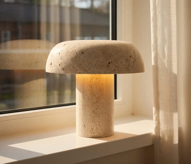 May include: A mushroom-shaped table lamp crafted from beige travertine stone with a porous, textured surface. The lamp features a wide, rounded dome shade resting on a cylindrical base, emitting a soft, warm glow from the center.