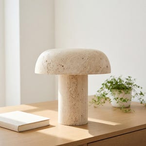 May include: A mushroom-shaped table lamp crafted from beige, porous travertine stone. The lamp features a wide, rounded dome shade resting on a cylindrical base, showcasing the natural, textured, and pitted surface of the stone material.