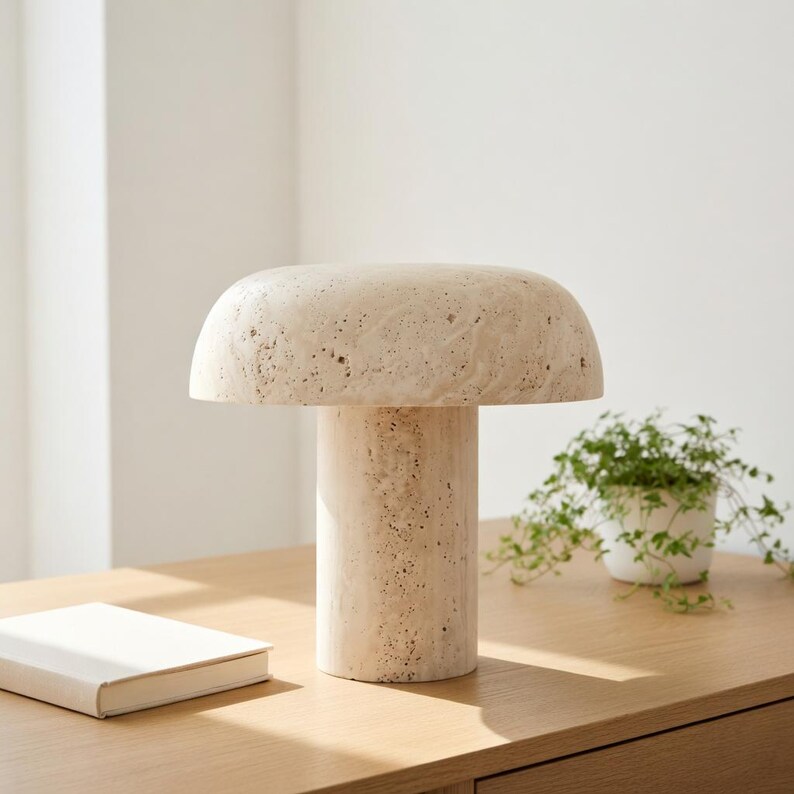May include: A mushroom-shaped table lamp crafted from beige, porous travertine stone. The lamp features a wide, rounded dome shade resting on a cylindrical base, showcasing the natural, textured, and pitted surface of the stone material.