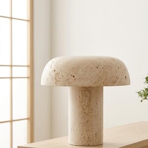 May include: A mushroom-shaped table lamp crafted from beige travertine stone. The lamp features a wide, rounded dome shade resting on a cylindrical base, both displaying the natural porous texture and cream-colored variations of the stone material.