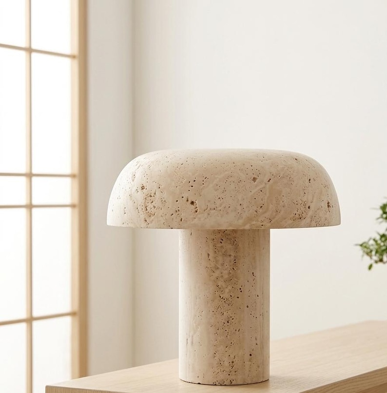 May include: A mushroom-shaped table lamp crafted from beige travertine stone. The lamp features a wide, rounded dome shade resting on a cylindrical base, both displaying the natural porous texture and cream-colored variations of the stone material.