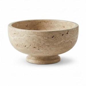 May include: A beige stone bowl with a pedestal base. The bowl has a rounded shape with a wide opening and a textured surface. The pedestal base is circular and supports the bowl. This decorative bowl is suitable for home decor.