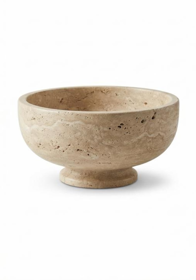 May include: A beige stone bowl with a pedestal base. The bowl has a rounded shape with a wide opening and a textured surface. The pedestal base is circular and supports the bowl. This decorative bowl is suitable for home decor.