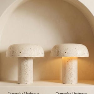 May include: Two mushroom-shaped lamps crafted from beige travertine stone with a porous, textured surface. One lamp is shown in the off position, while the other is illuminated, casting a warm glow from beneath its wide, rounded cap.