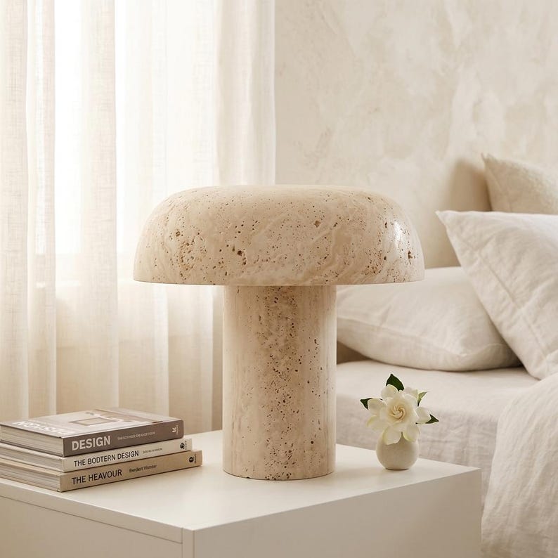 May include: A mushroom-shaped table lamp crafted from beige, porous travertine stone. The lamp features a wide, rounded dome shade resting on a cylindrical base, showcasing the natural, textured, and pitted surface of the stone material.