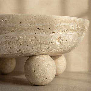 May include: A beige, textured stone bowl with a rounded shape, supported by three spherical feet. The bowl has a wide, shallow design and a natural, earthy aesthetic. The stone has visible variations in color and texture.
