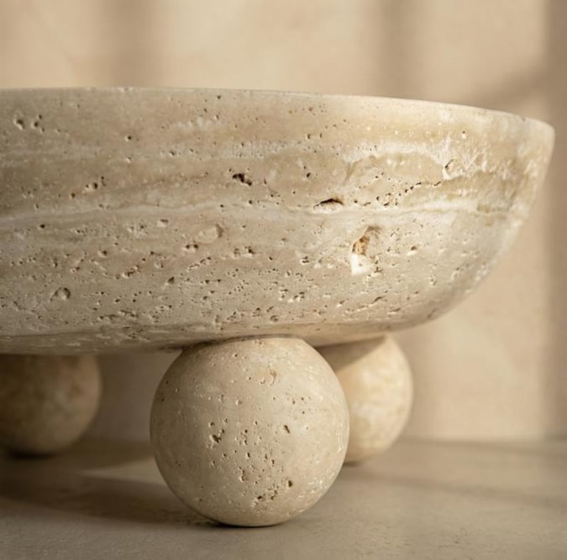 May include: A beige, textured stone bowl with a rounded shape, supported by three spherical feet. The bowl has a wide, shallow design and a natural, earthy aesthetic. The stone has visible variations in color and texture.