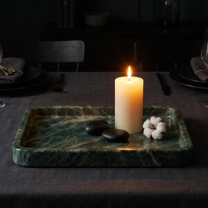 May include: A rectangular green marble tray holds a lit white candle, two black stones, and a cotton flower. The tray sits on a dark grey tablecloth, creating a serene and elegant ambiance.