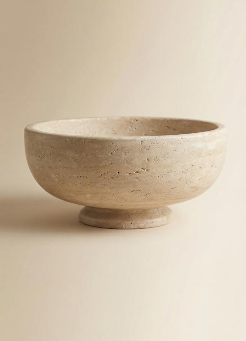 May include: A beige stone bowl with a rounded shape and a pedestal base. The bowl has a textured surface and a wide opening. The neutral colour and simple design make it a versatile decorative piece.