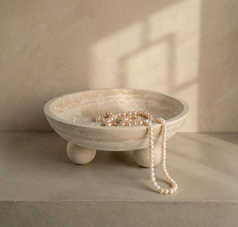May include: A beige stone bowl with three rounded feet, containing a strand of pearls. The bowl has a smooth, rounded shape and a natural, textured appearance. The pearls are a soft, creamy colour, and the bowl sits on a neutral surface.
