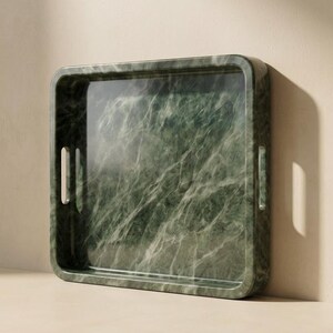 May include: A square, green marble tray with a polished finish. The tray has a rectangular interior and rounded corners. Two vertical handle cutouts are on opposite sides. The marble has a natural veining pattern.