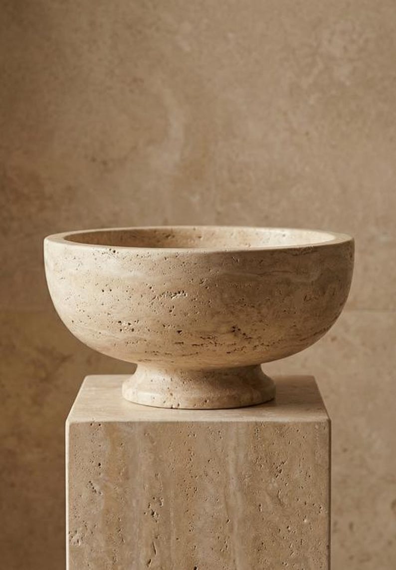 May include: A beige stone bowl with a rounded form, placed on a matching square plinth. The bowl features a wide opening and a textured surface. The plinth is also made of stone and has a textured surface.