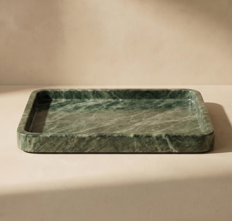 May include: A rectangular tray crafted from green marble. The tray features rounded corners and a subtly raised edge. The marble displays a natural veining pattern, with green and white hues. The tray is placed on a light-coloured surface.