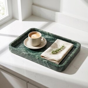 May include: A rectangular green marble tray holds a beige cup and saucer with coffee, and a folded white napkin with a sprig of rosemary. The tray sits on a white countertop near a window.