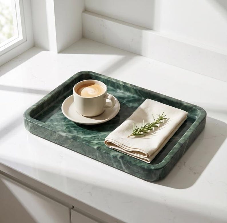 May include: A rectangular green marble tray holds a beige cup and saucer with coffee, and a folded white napkin with a sprig of rosemary. The tray sits on a white countertop near a window.