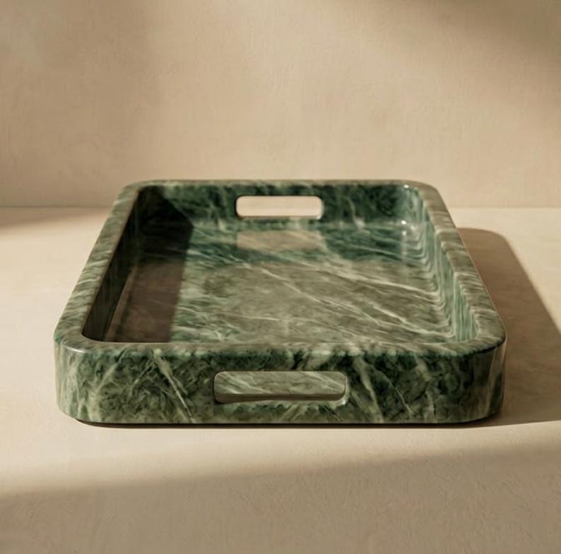 May include: A rectangular green marble tray with integrated handles. The tray has a polished finish and a natural veining pattern. The handles are rectangular cutouts on the sides. A decorative item, the tray is made of marble.