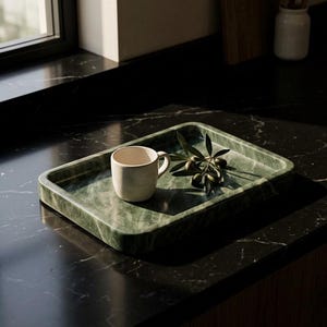May include: A green marble tray with a rounded rectangular shape. A small, off-white mug sits on the tray, along with a sprig of olives. The tray is on a dark countertop, with a window in the background.