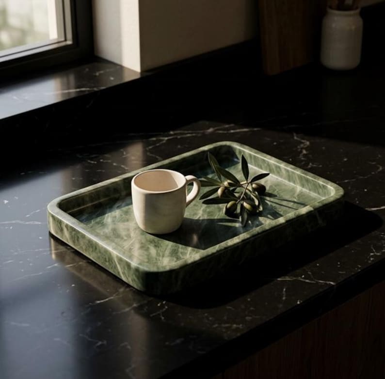 May include: A green marble tray with a rounded rectangular shape. A small, off-white mug sits on the tray, along with a sprig of olives. The tray is on a dark countertop, with a window in the background.
