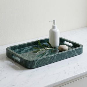 May include: A rectangular, dark green marble tray with integrated handles. A white soap dispenser, a small stone, and a sprig of greenery rest on the tray. The tray is on a white marble surface.