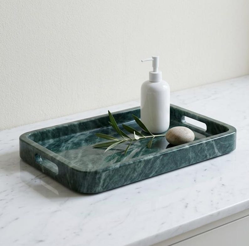 May include: A rectangular, dark green marble tray with integrated handles. A white soap dispenser, a small stone, and a sprig of greenery rest on the tray. The tray is on a white marble surface.