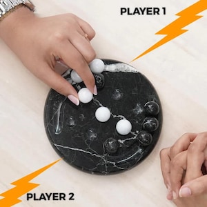May include: A marble board game with black and white marbles on a round, black and white marble patterned board. The text "PLAYER 1" and "PLAYER 2" are visible. Two people are playing the game.