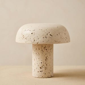 May include: A mushroom-shaped table lamp crafted from beige travertine stone. The lamp features a smooth, rounded cap and a cylindrical base, both displaying the natural, porous, and pitted texture characteristic of raw, unpolished travertine stone.