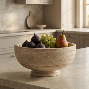May include: A beige stone fruit bowl filled with fresh figs, green grapes, and a pear. The bowl sits on a light-coloured countertop in a kitchen setting. The bowl has a pedestal base.