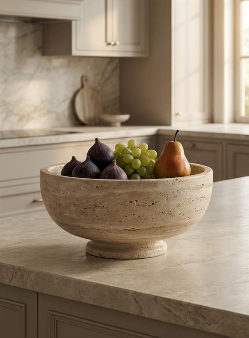 May include: A beige stone fruit bowl filled with fresh figs, green grapes, and a pear. The bowl sits on a light-coloured countertop in a kitchen setting. The bowl has a pedestal base.
