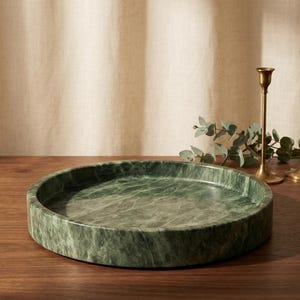 May include: A circular, green marble tray with a natural, veined pattern. The tray is on a wooden surface. Two gold candle holders and greenery are in the background, suggesting a decorative or serving piece.