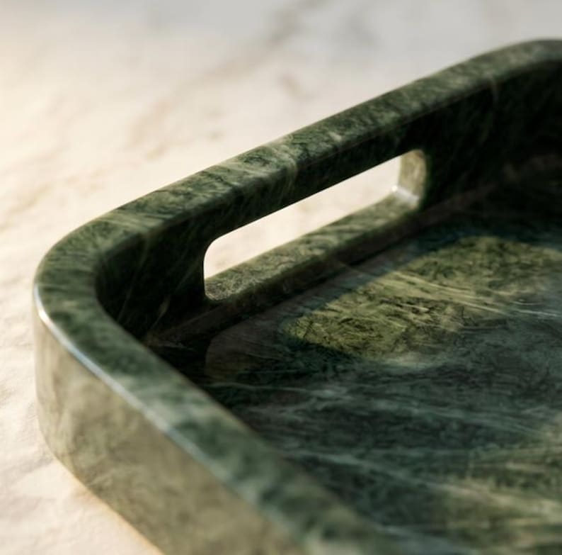 May include: Close-up of a rectangular, dark green marble tray with rounded corners and a handle cut-out. The tray's surface has a natural, veined pattern. The tray is designed for serving or display.