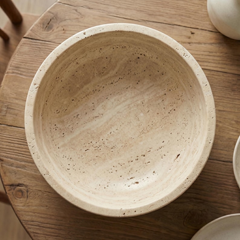 May include: A beige, round bowl with a textured surface. The bowl is made of a light-coloured stone material and sits on a wooden table. The bowl's interior is smooth, and the exterior has a slightly rough texture.