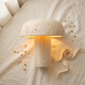 May include: A mushroom-shaped table lamp crafted from beige porous stone with a textured surface. The lamp features a wide, rounded cap and a cylindrical base, emitting a warm, soft glow from beneath the cap, illuminating the surrounding area.
