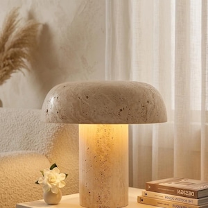 May include: A mushroom-shaped table lamp crafted from beige travertine stone with a porous, textured surface. The lamp features a wide, rounded shade resting on a cylindrical base, emitting a warm, soft glow from the center of the structure.