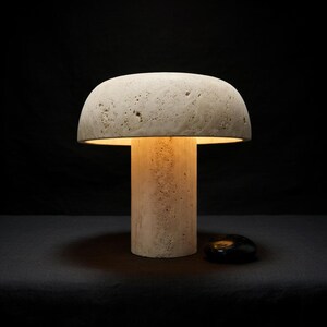 May include: A mushroom-shaped table lamp crafted from beige, porous stone with a textured surface. The lamp features a cylindrical base and a wide, domed shade that emits a warm, soft glow from underneath, illuminating the stone's natural cavities.