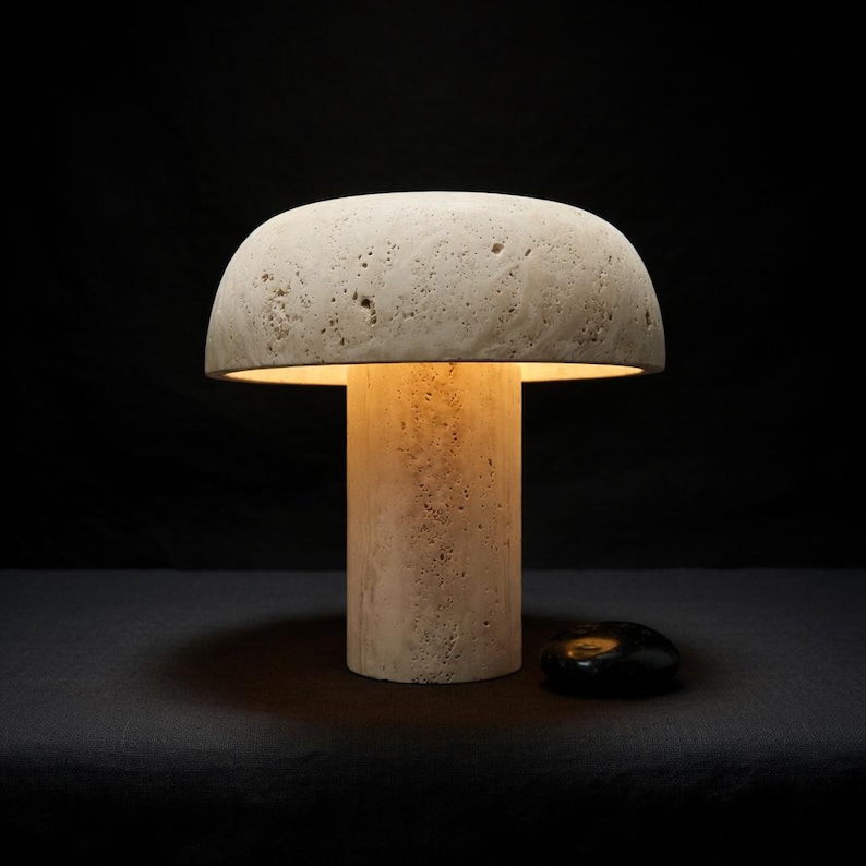 May include: A mushroom-shaped table lamp crafted from beige, porous stone with a textured surface. The lamp features a cylindrical base and a wide, domed shade that emits a warm, soft glow from underneath, illuminating the stone's natural cavities.