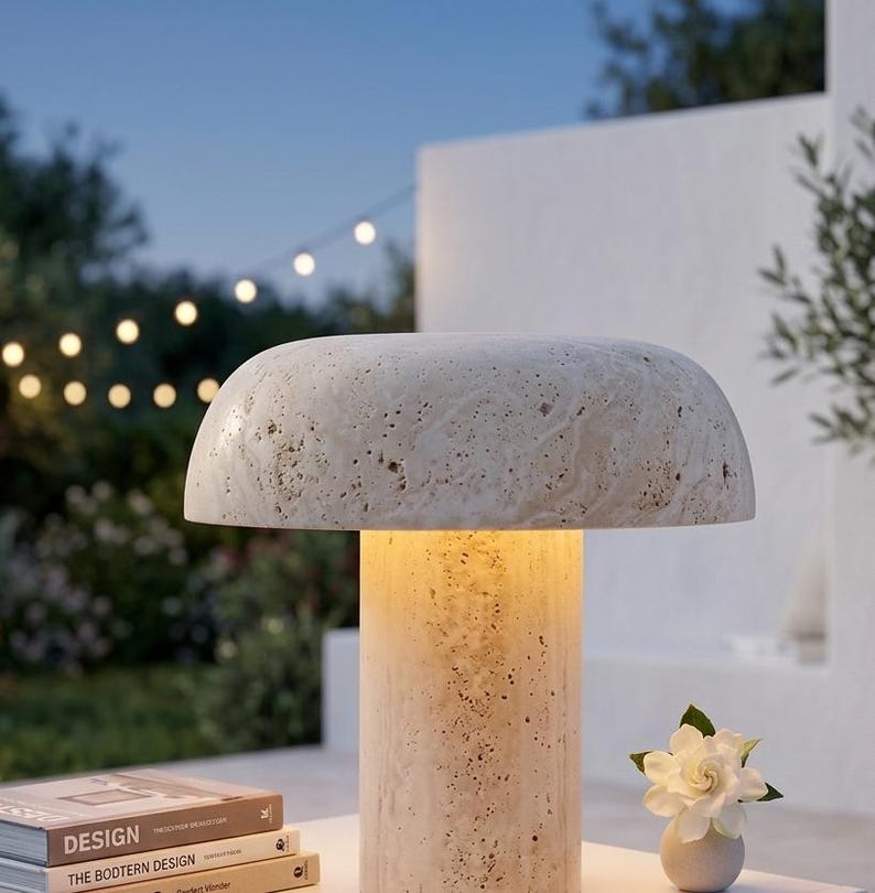 May include: A mushroom-shaped table lamp crafted from beige travertine stone with a porous, textured surface. The lamp features a wide, rounded shade resting on a cylindrical base, emitting a warm, soft glow from the center of the structure.